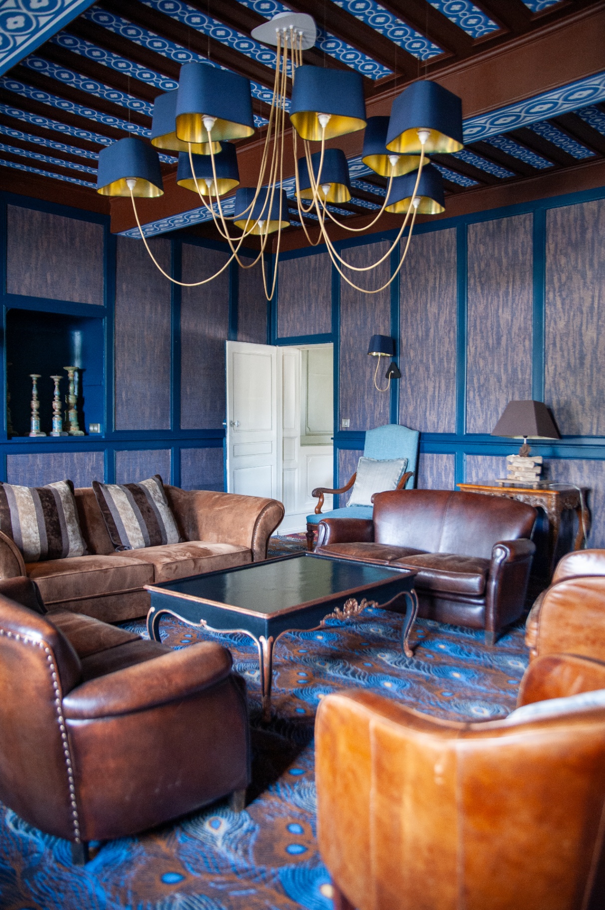 Salon bleu lambrissé avec plafond à caissons, lustre doré et fauteuils club en cuir