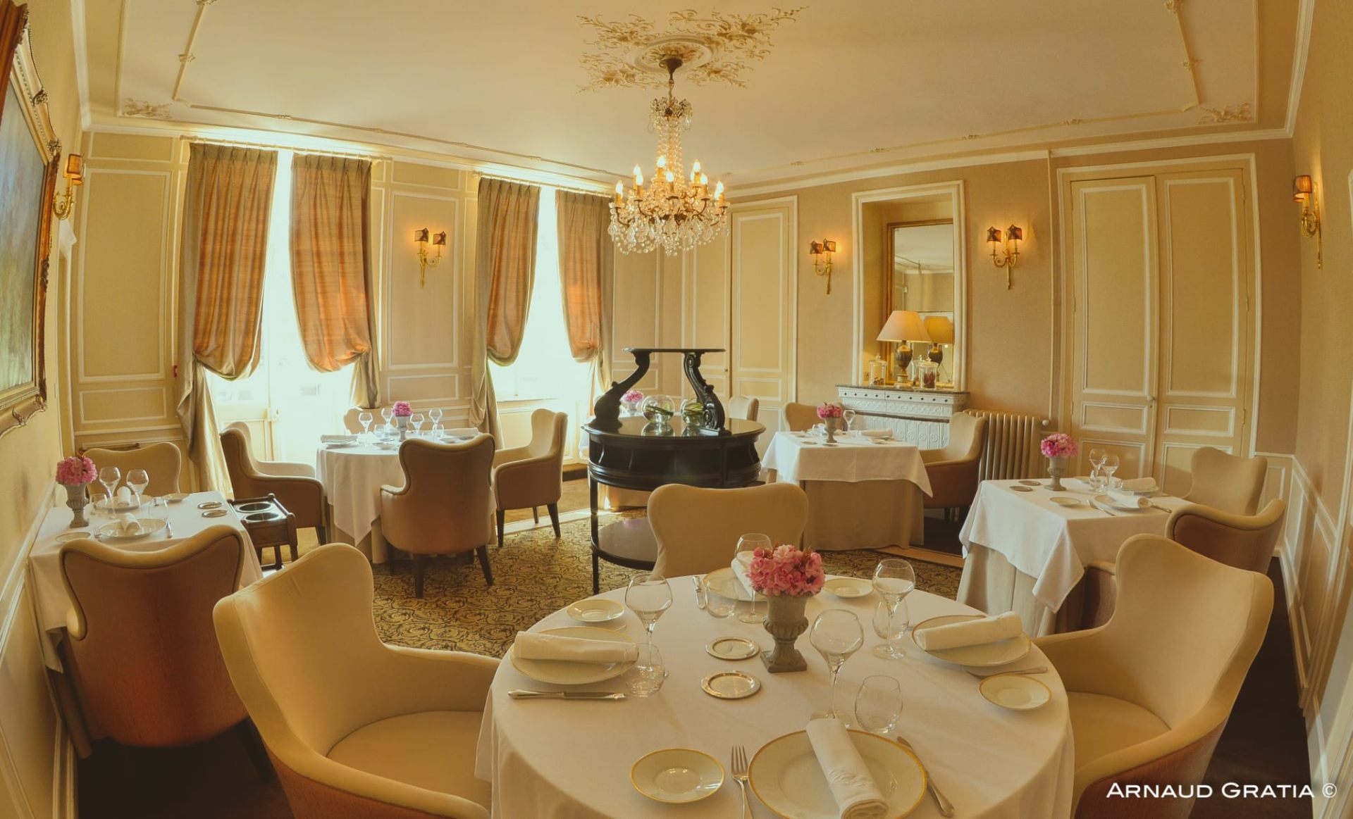 Salle de restaurant aux boiseries crème, lustre en cristal et tables nappées de blanc avec compositions florales roses